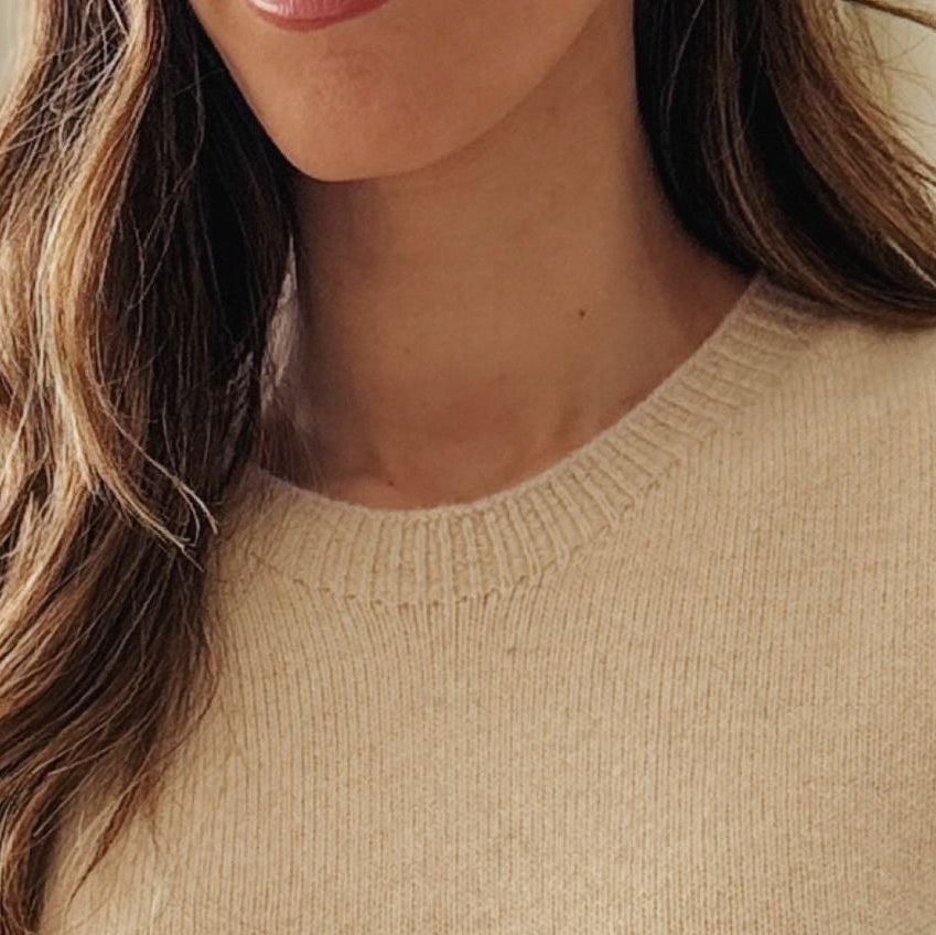 A close-up of a woman wearing a soft cream-colored knit sweater, with her hair and lower face visible against a bright indoor background.