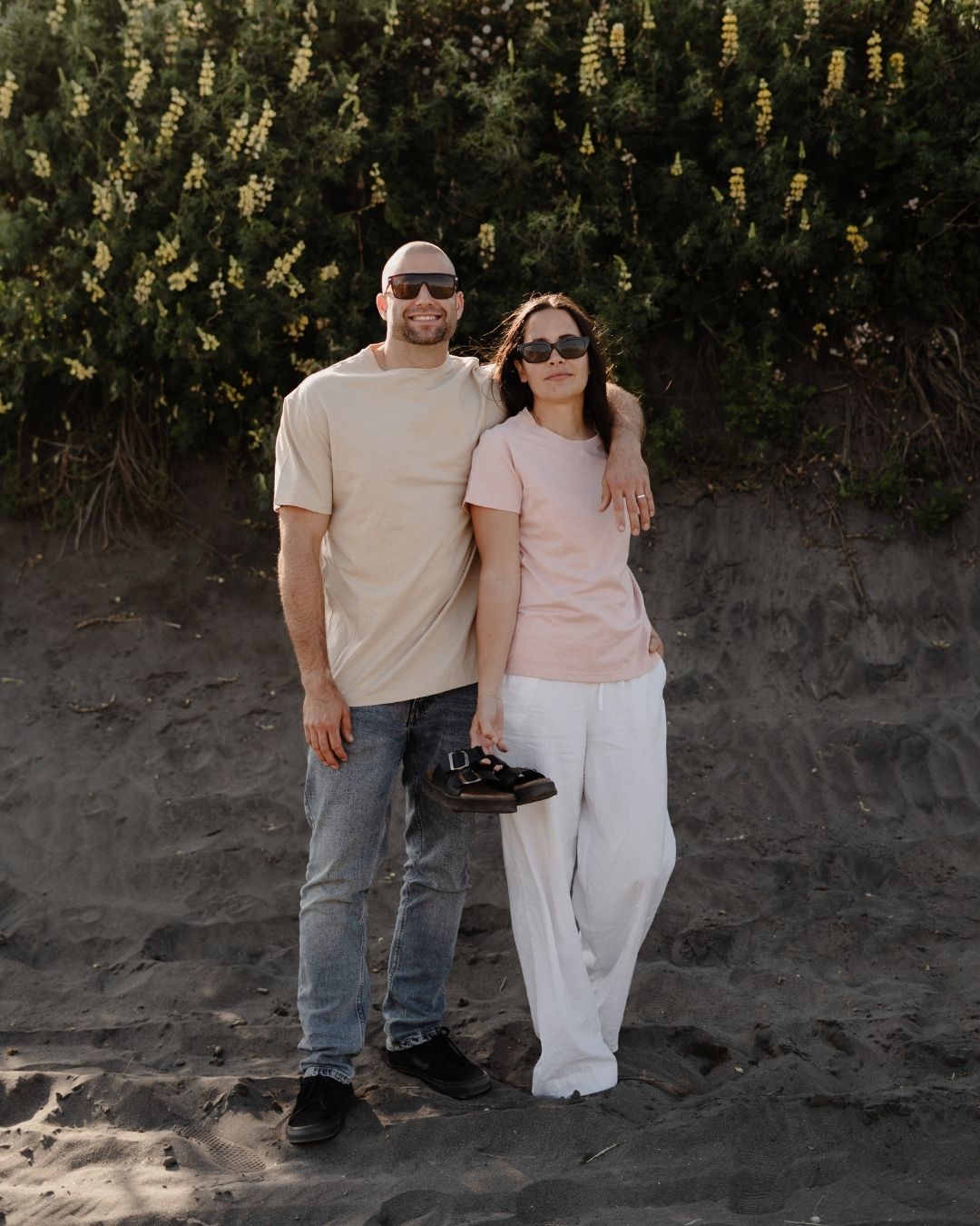 Two people standing on a sandy beach with greenery in the background