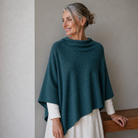 Woman wearing a teal poncho over a white dress, standing against a plain background